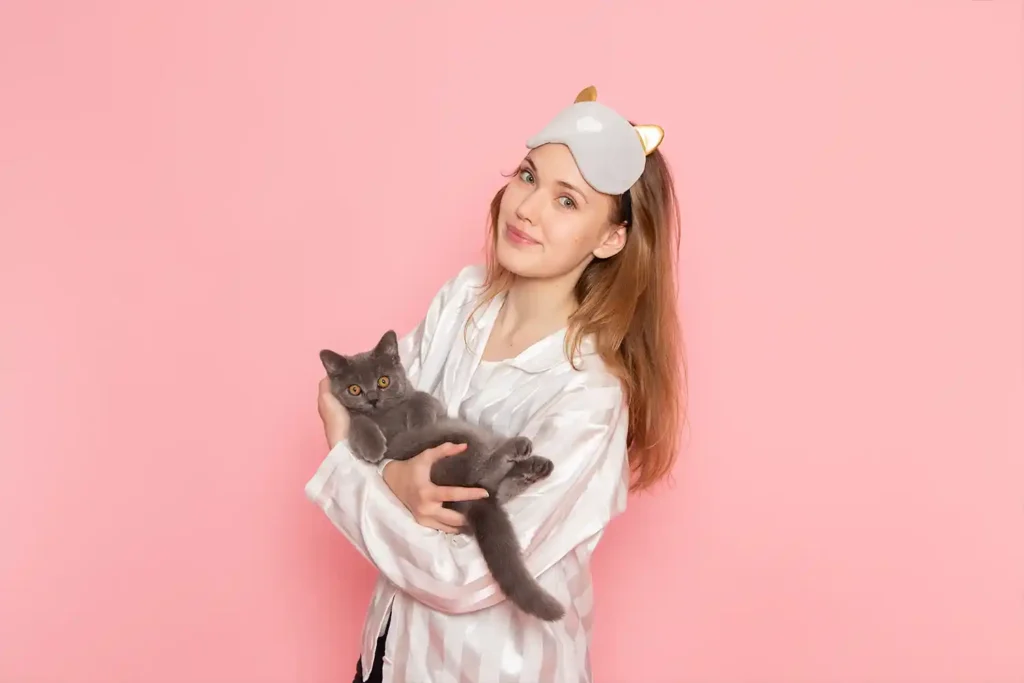 Russian Blue cat lying comfortably on its back, held by a woman in pajamas with a cat sleep cover on its forehead, both looking at the camera against a pink studio background.